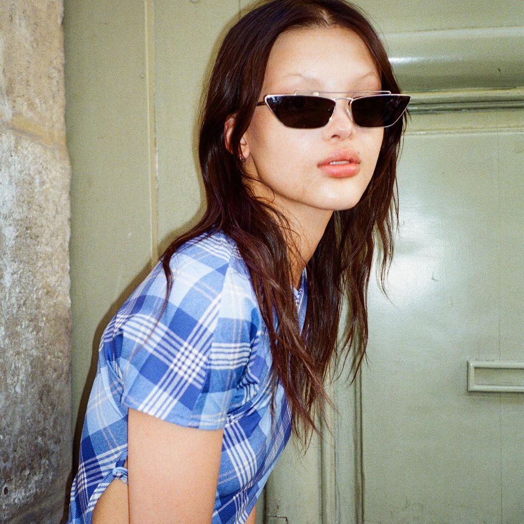 Portrait of a woman with long brown hair wearing sunglasses and a blue plaid shirt, standing in front of a green door.