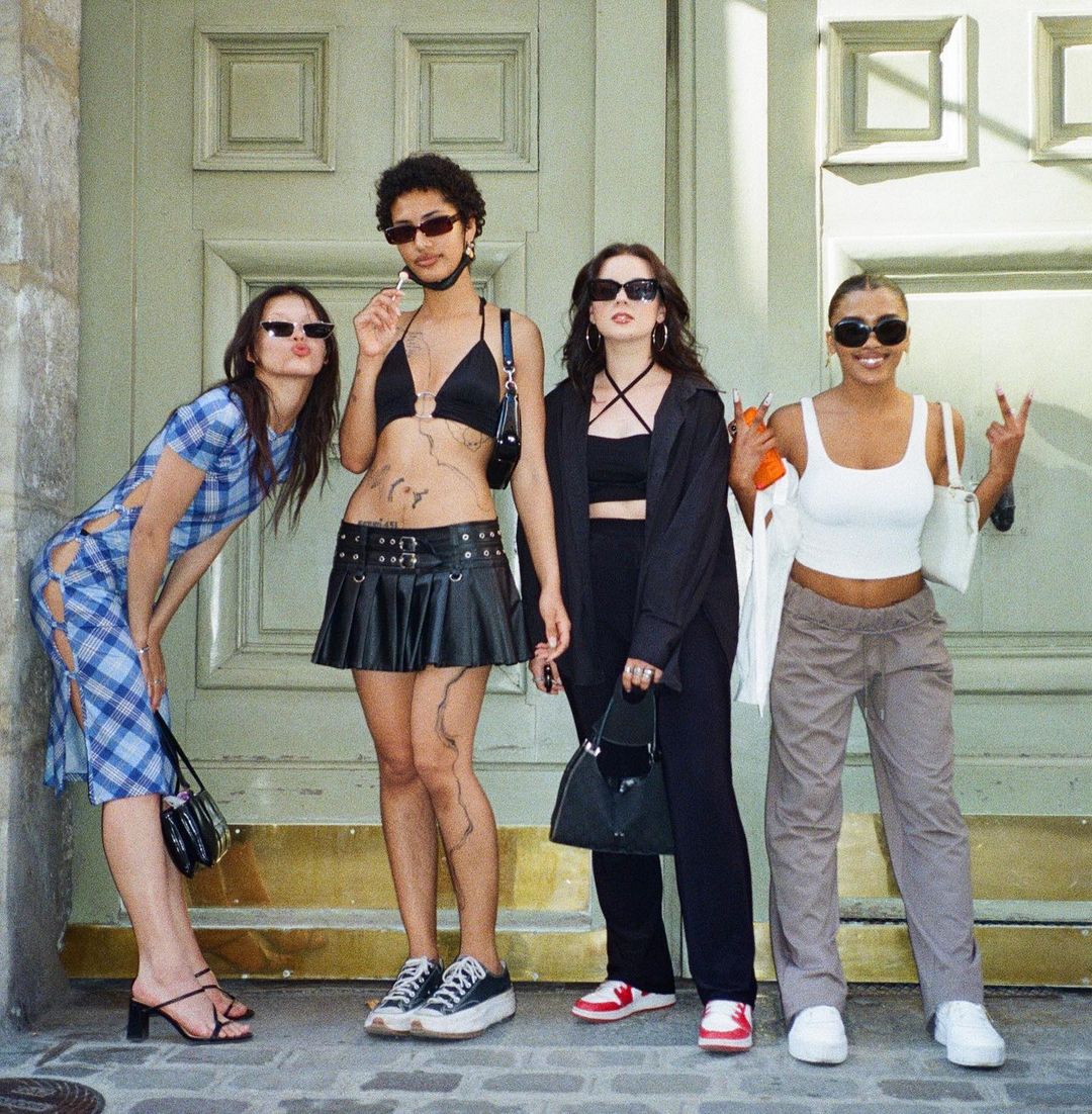 Four young women pose in front of a large, light-green door, displaying diverse casual summer fashion with sunglasses and purses.