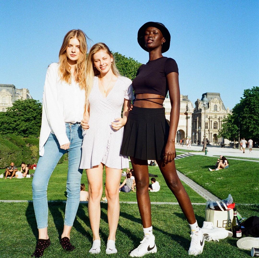 Three women pose in a park with European architecture, wearing casual dresses and tops, under a clear blue sky on a sunny day.