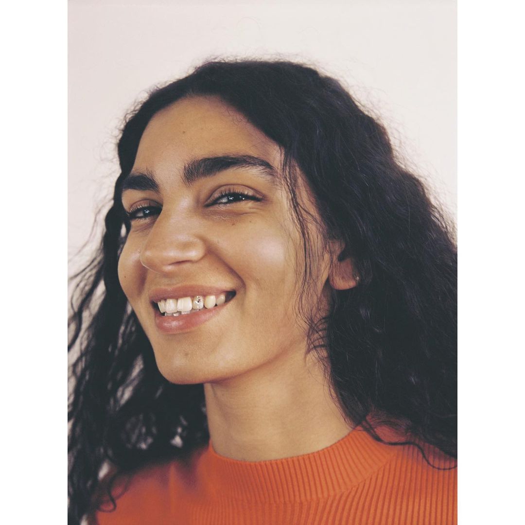 Portrait of a smiling woman with long, dark wavy hair wearing an orange top against a neutral background. Natural light.