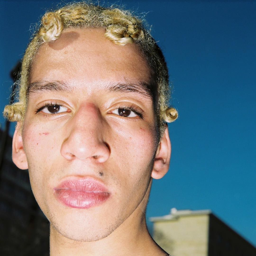 Close-up portrait of a young adult with light skin, short blonde hair, and brown eyes set against a blue sky and buildings.