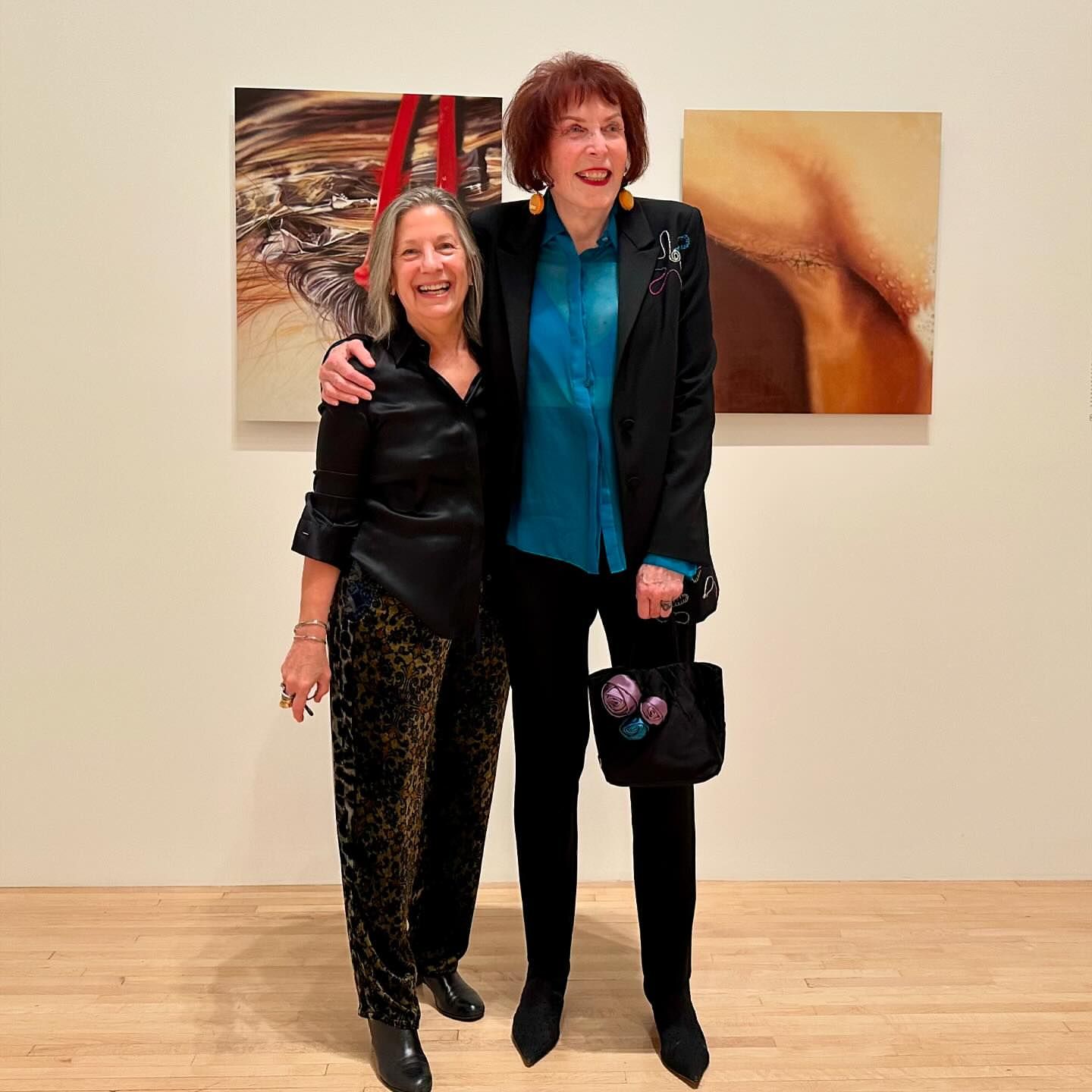 Two women pose in front of abstract paintings in an art gallery. One woman has shoulder length hair and animal print pants.