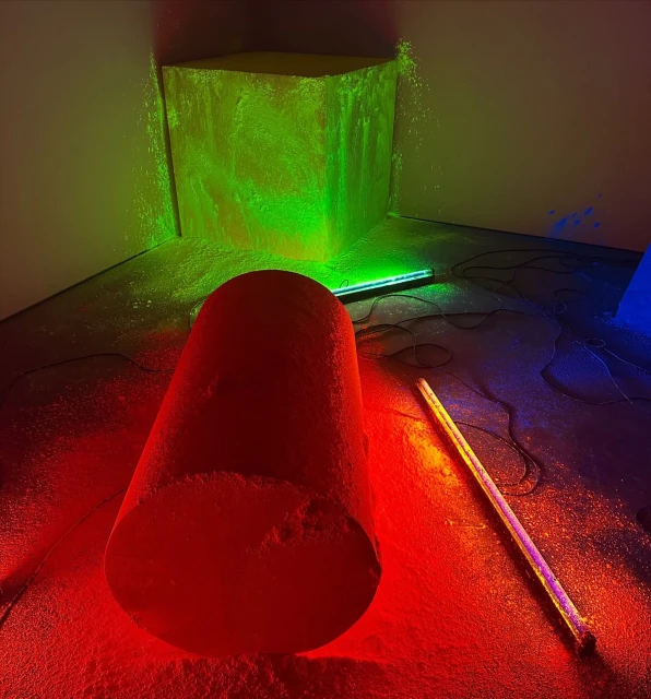 Abstract art installation featuring a red cylinder, green cube, and blue element in a white room with colorful lighting and dark flooring.