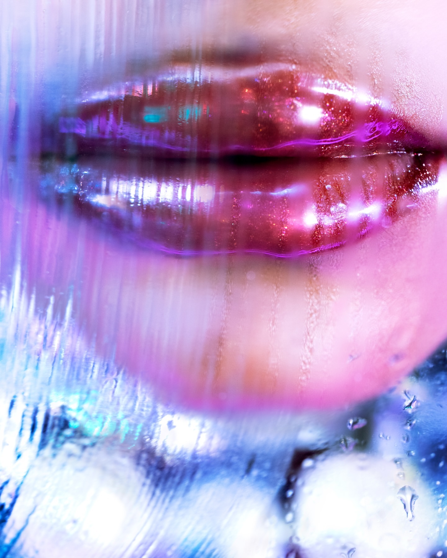 Close-up of glistening lips with pink gloss and purple outline, blurred by water droplets on glass in a vibrant, artistic beauty shot.