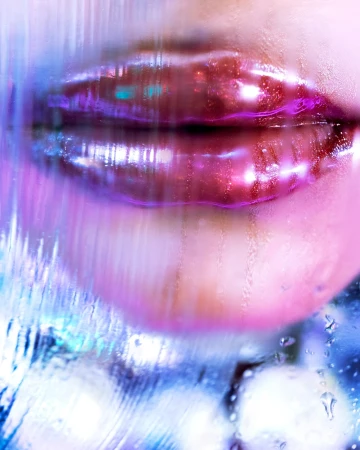 Close-up of glistening lips with pink gloss and purple outline, blurred by water droplets on glass in a vibrant, artistic beauty shot.