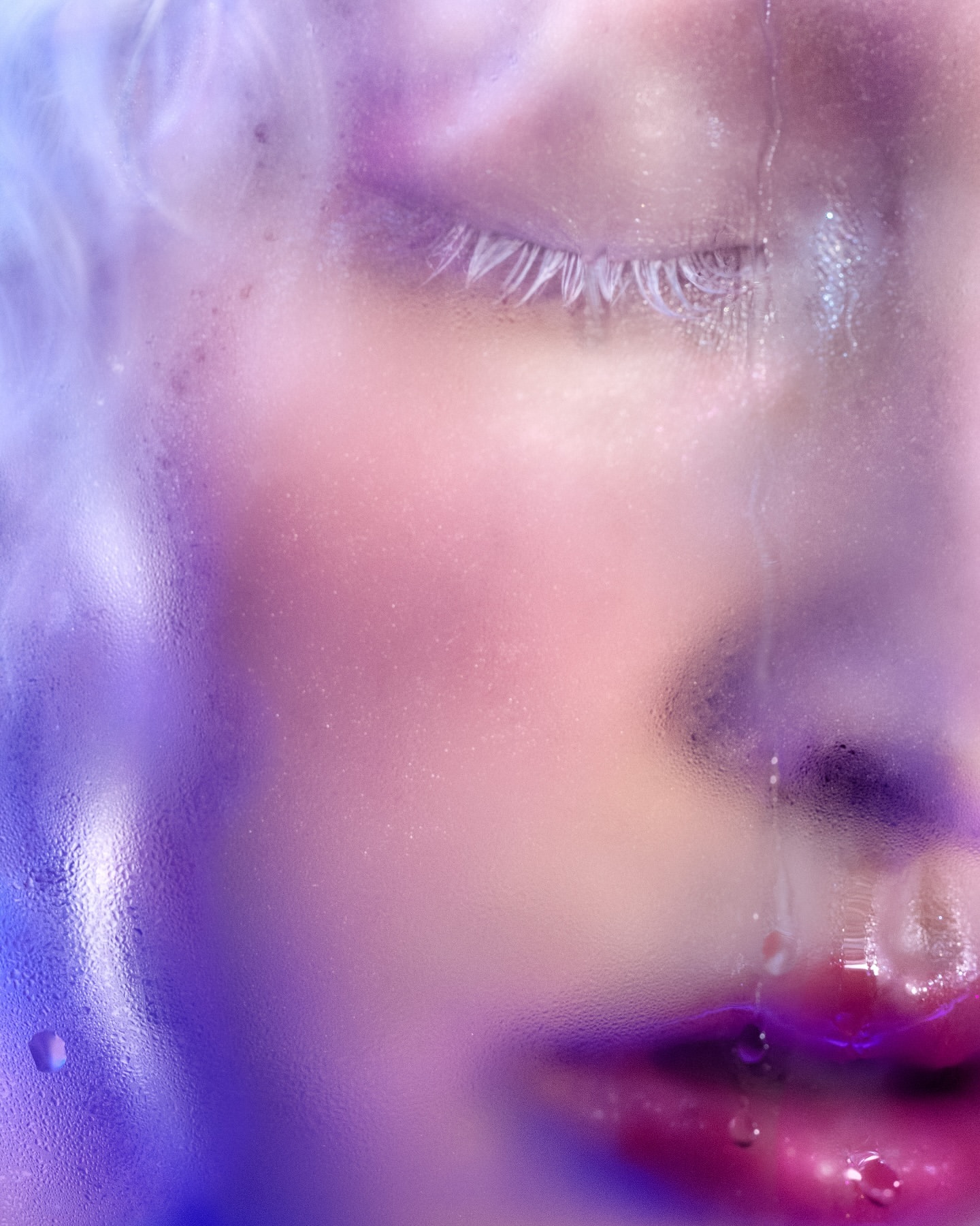 Close-up portrait of a face with pink lips, closed eyes, and water droplets on textured glass creating a dreamy, ethereal effect.