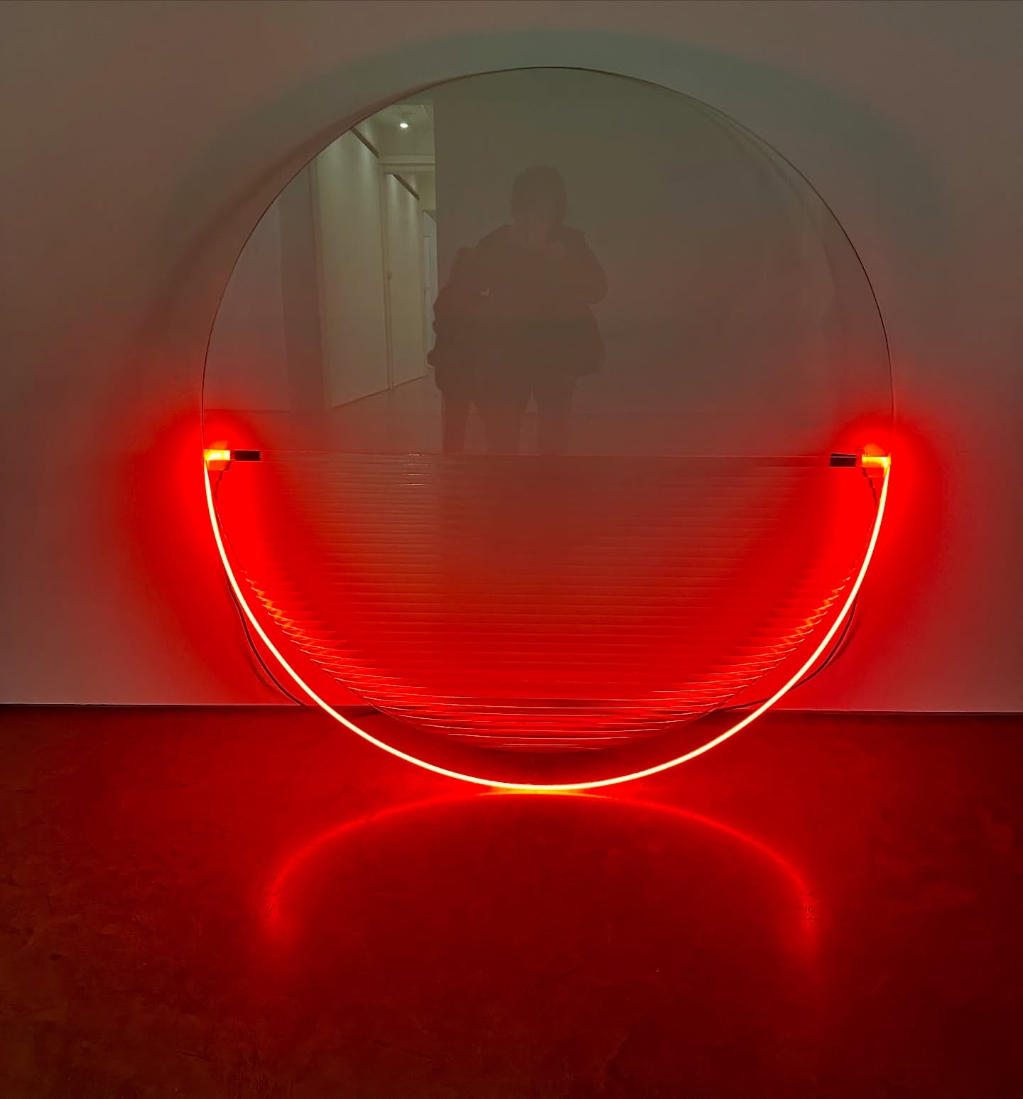 Abstract art installation featuring a round mirror with a red, semi-circular neon light fixture against a white wall in an indoor setting.