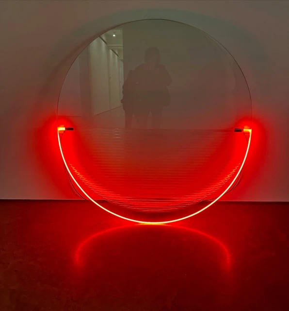 Abstract art installation featuring a round mirror with a red, semi-circular neon light fixture against a white wall in an indoor setting.