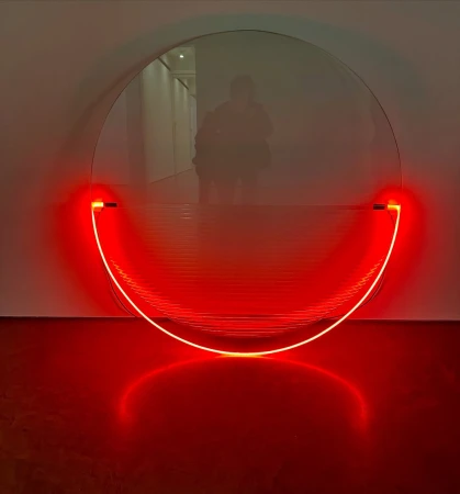 Abstract art installation featuring a round mirror with a red, semi-circular neon light fixture against a white wall in an indoor setting.