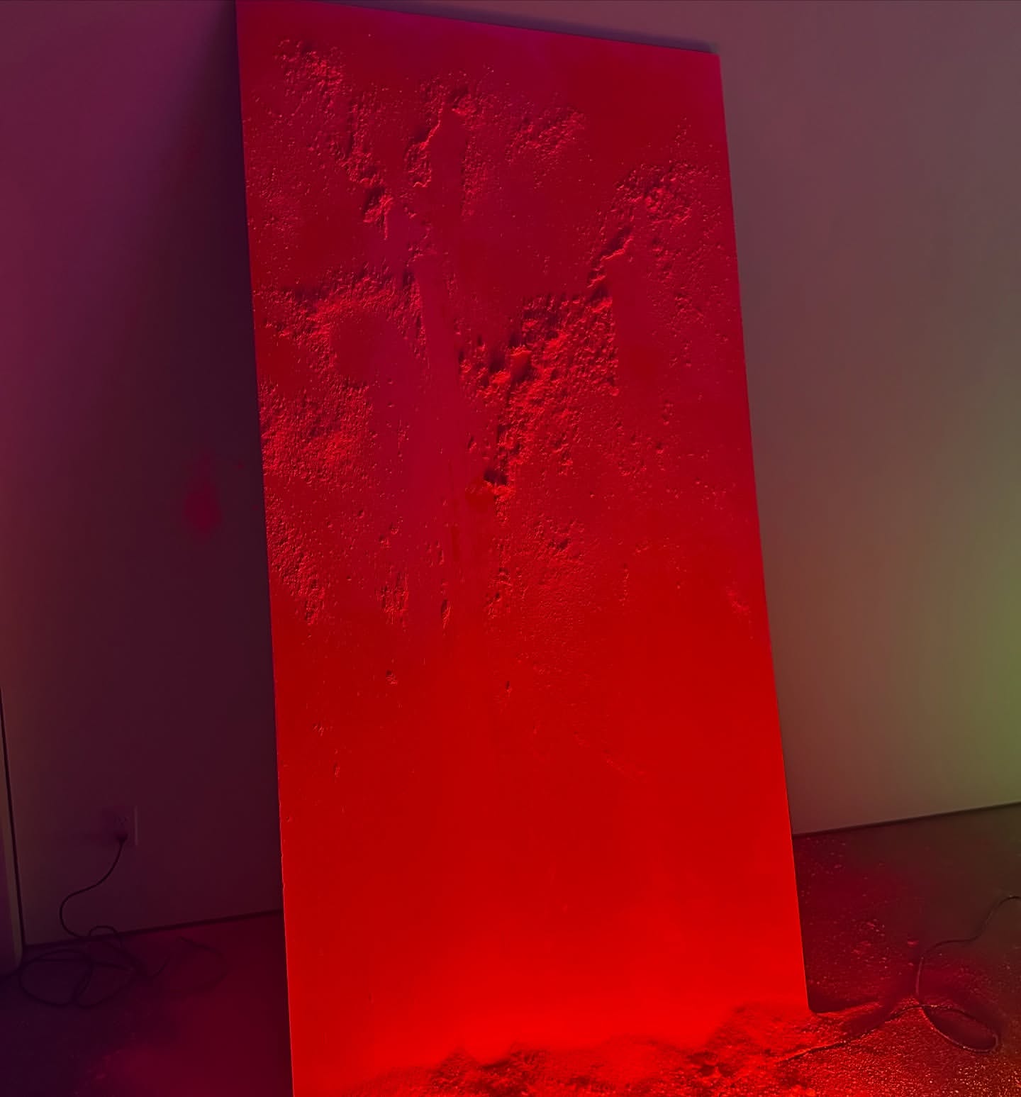 Abstract art piece in red and pink hues featuring a textured surface leaning against a wall in a dimly lit indoor setting.