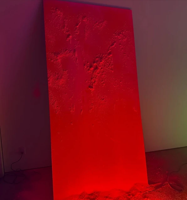 Abstract art piece in red and pink hues featuring a textured surface leaning against a wall in a dimly lit indoor setting.