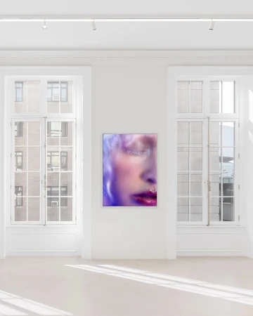 Framed photographic artwork depicting a close-up portrait of a woman with vibrant makeup hung on a white wall between two large windows.
