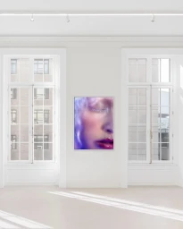 Framed photographic artwork depicting a close-up portrait of a woman with vibrant makeup hung on a white wall between two large windows.