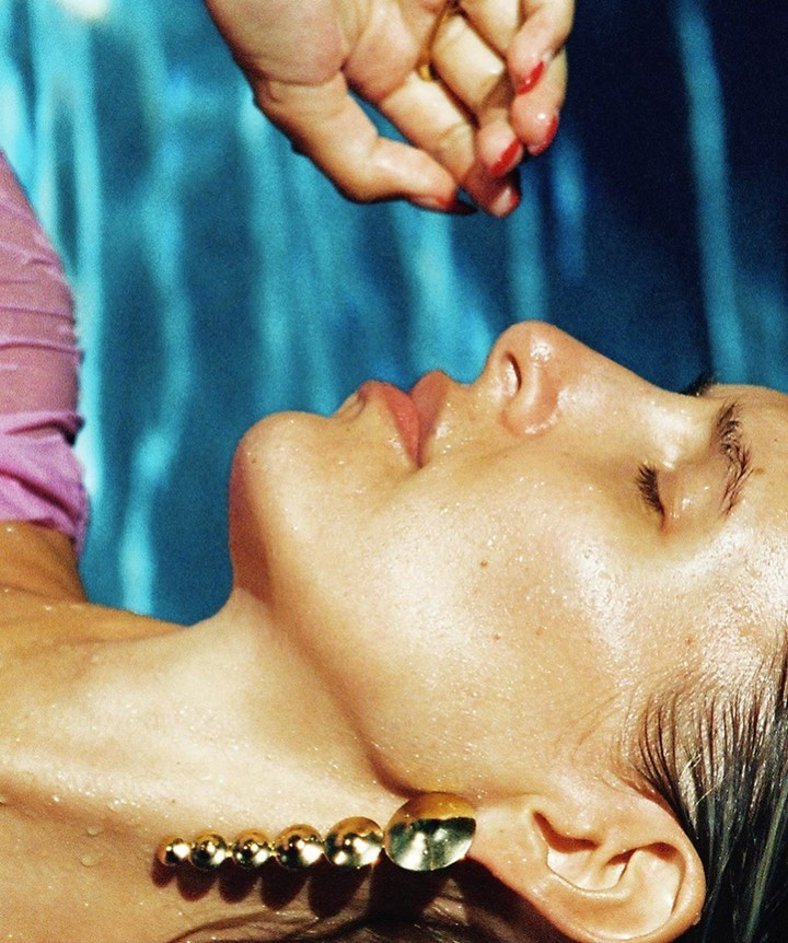 Close-up view of a woman lying back in a pool of water, with another hand above her, and wearing a gold earring and pink top.
