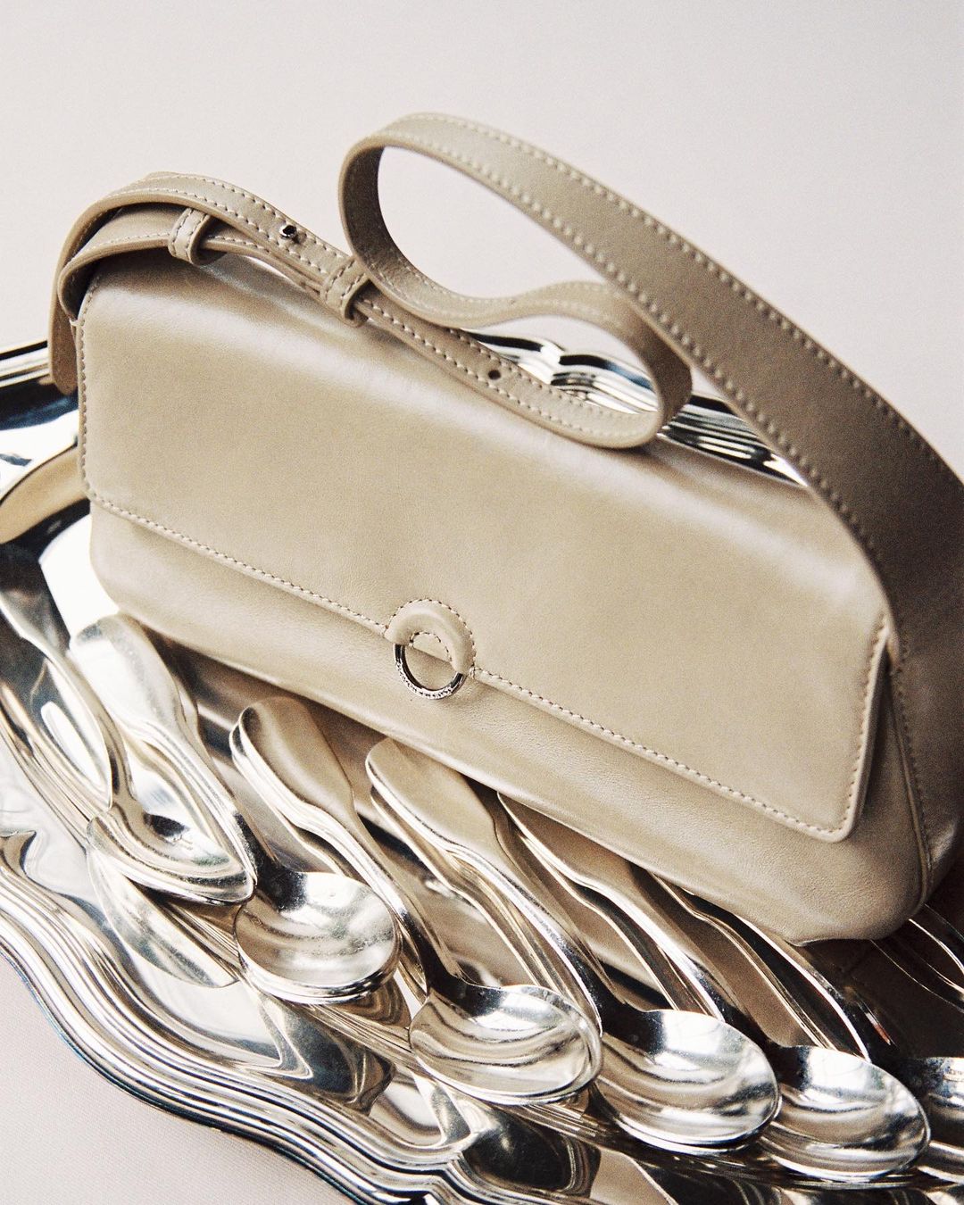 Elegant beige leather handbag with a shoulder strap placed on top of an ornate silver platter filled with vintage silver spoons.