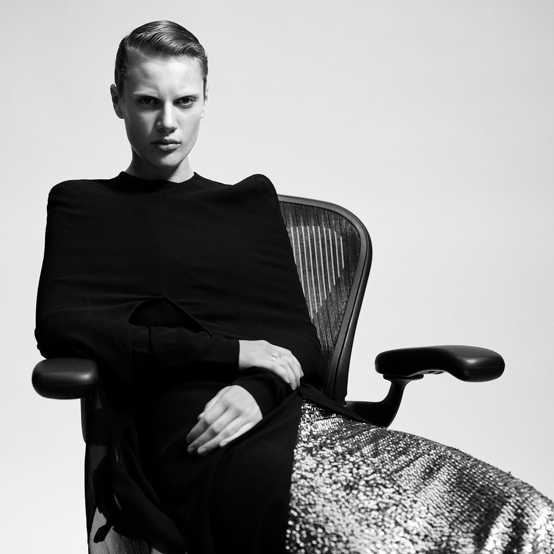 Monochrome portrait of a poised woman in an office chair, wearing a black top and glittering skirt, set against a neutral background.