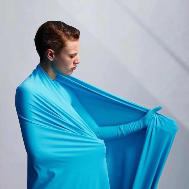 A stylish young male model posing indoors while wrapped in draped blue fabric, with a side profile view and clean studio lighting.