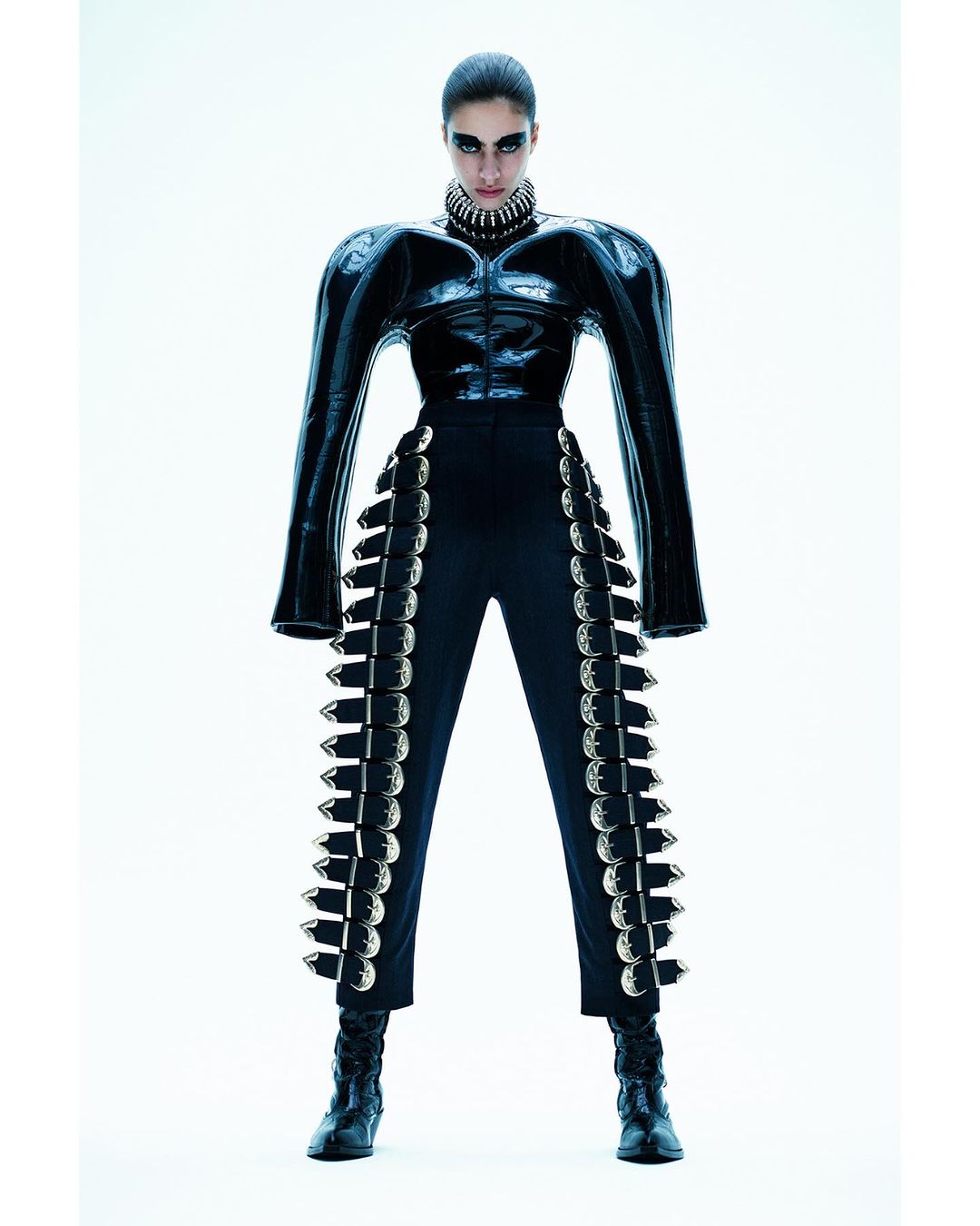Fashion shot of a woman in a black, futuristic, studded outfit standing in a bright white photo studio, looking at the camera with a serious expression.