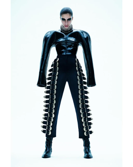 Fashion shot of a woman in a black, futuristic, studded outfit standing in a bright white photo studio, looking at the camera with a serious expression.
