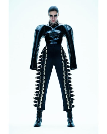 Fashion shot of a woman in a black, futuristic, studded outfit standing in a bright white photo studio, looking at the camera with a serious expression.