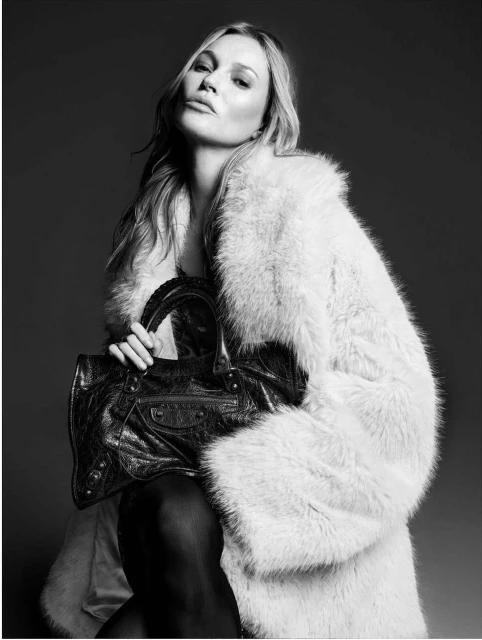 Blonde woman in a fur coat poses indoors with a black handbag, creating a high-contrast fashion portrait against a dark background.