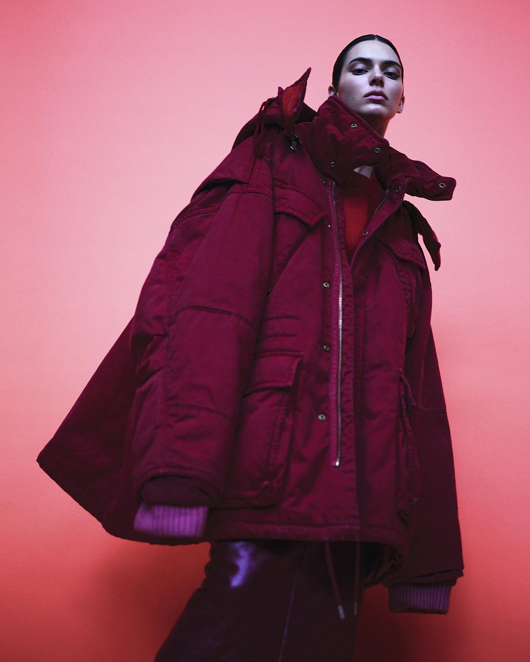 Fashion shot of a young woman in a red puffer coat and black pants on a pink background, looking at the camera in a studio setting.