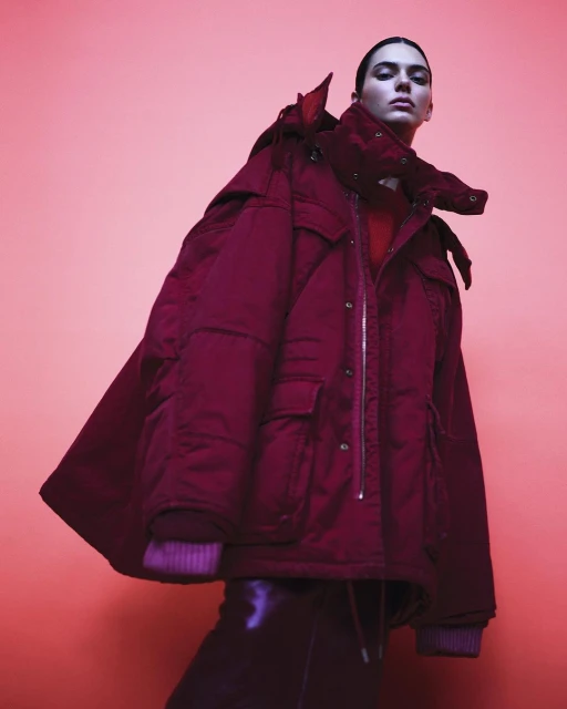 Fashion shot of a young woman in a red puffer coat and black pants on a pink background, looking at the camera in a studio setting.