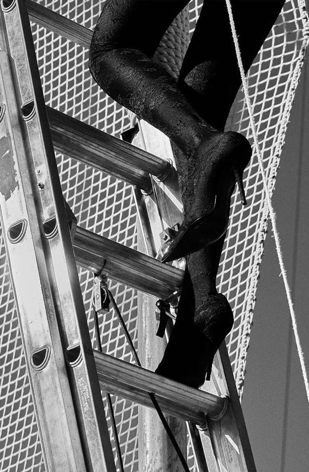 Close-up black and white shot of woman's legs and feet in black high heels climbing a metal ladder against a mesh background.