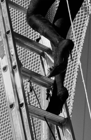 Close-up black and white shot of woman's legs and feet in black high heels climbing a metal ladder against a mesh background.