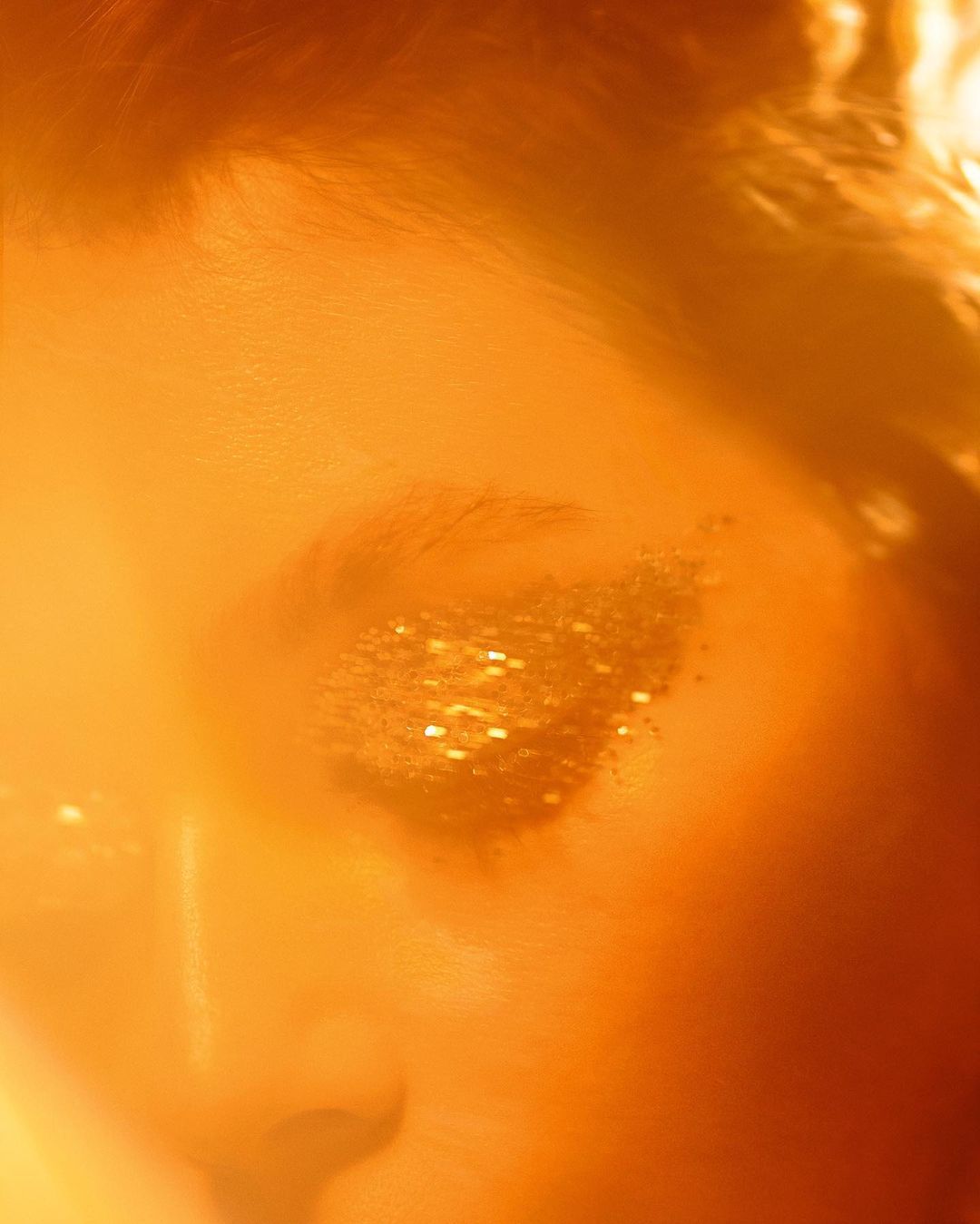 Close-up of a woman's face with glittering gold eyeshadow makeup, shot with warm light and shallow depth of field.