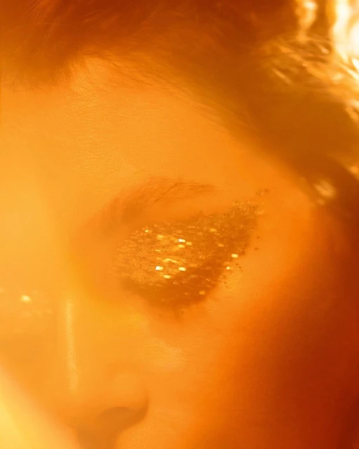 Close-up of a woman's face with glittering gold eyeshadow makeup, shot with warm light and shallow depth of field.