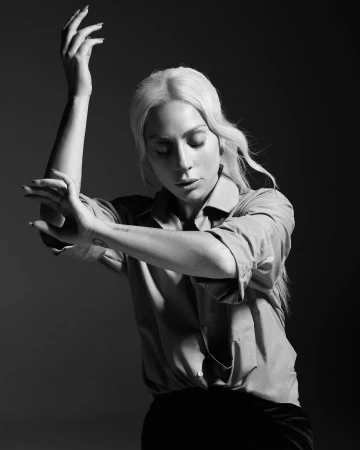 Monochrome portrait of Lady Gaga with blonde hair wearing a collared shirt, posing with arms raised in a studio setting by Mario Sorrenti.
