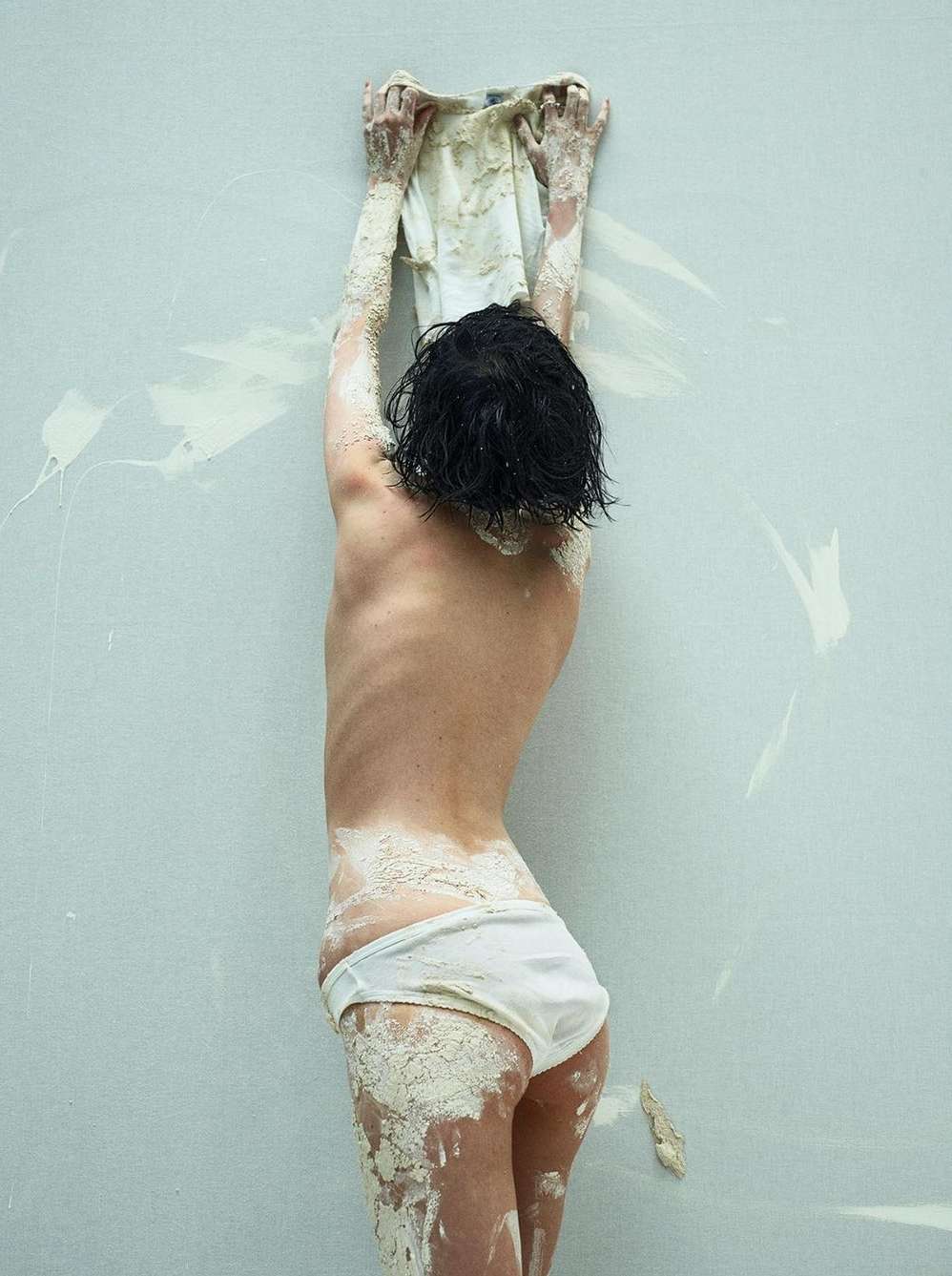 Rear view of a woman with dark wet hair wearing white underwear covered in pale mud reaching up against a concrete wall