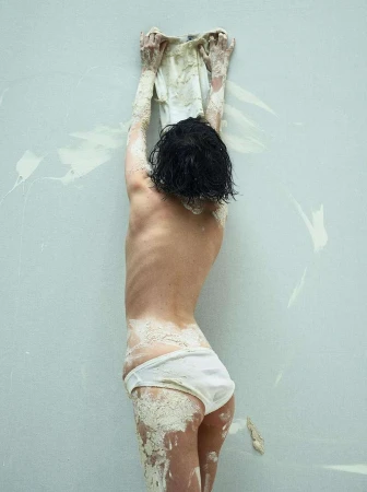 Rear view of a woman with dark wet hair wearing white underwear covered in pale mud reaching up against a concrete wall