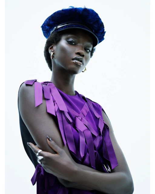 Fashion portrait of a black woman wearing a purple ribbon dress and a navy blue cap, striking a pose against a white backdrop.