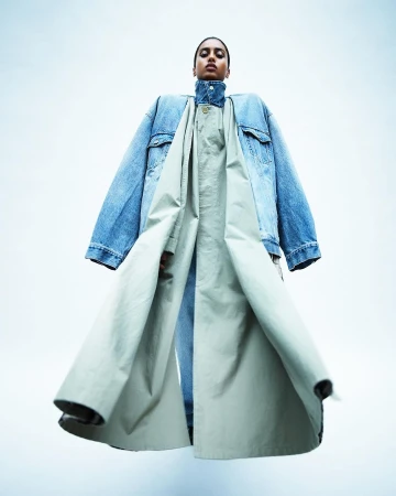 Low-angle full-length shot of a model wearing an oversized denim jacket and a long sage trench coat against a white background.