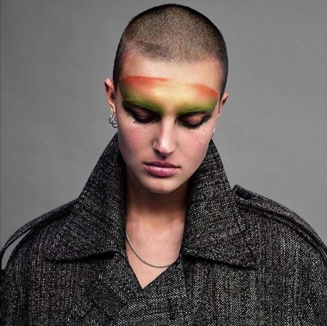 Close-up portrait of a woman with a shaved head wearing bold eye makeup and a grey coat, looking down against a grey background.
