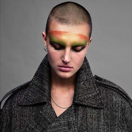 Close-up portrait of a woman with a shaved head wearing bold eye makeup and a grey coat, looking down against a grey background.
