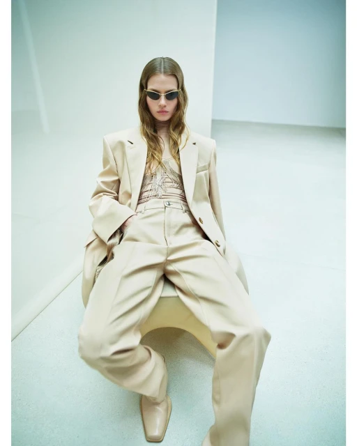 A stylish woman sits on a stool wearing a beige suit and sunglasses, captured in an indoor studio setting with natural lighting.