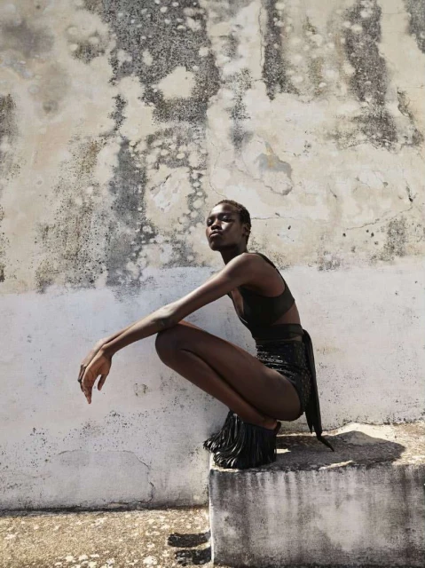 Woman in black, high-waisted shorts and sports bra, crouches on a concrete step in front of a textured, distressed wall, bathed in soft light.