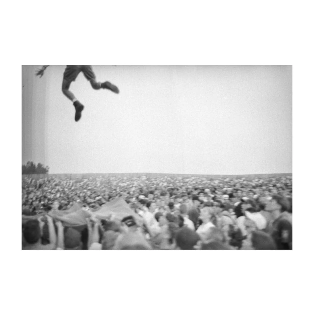 Monochrome image of an adult male jumping above a large crowd of people at an outdoor event, captured with a wide shot.