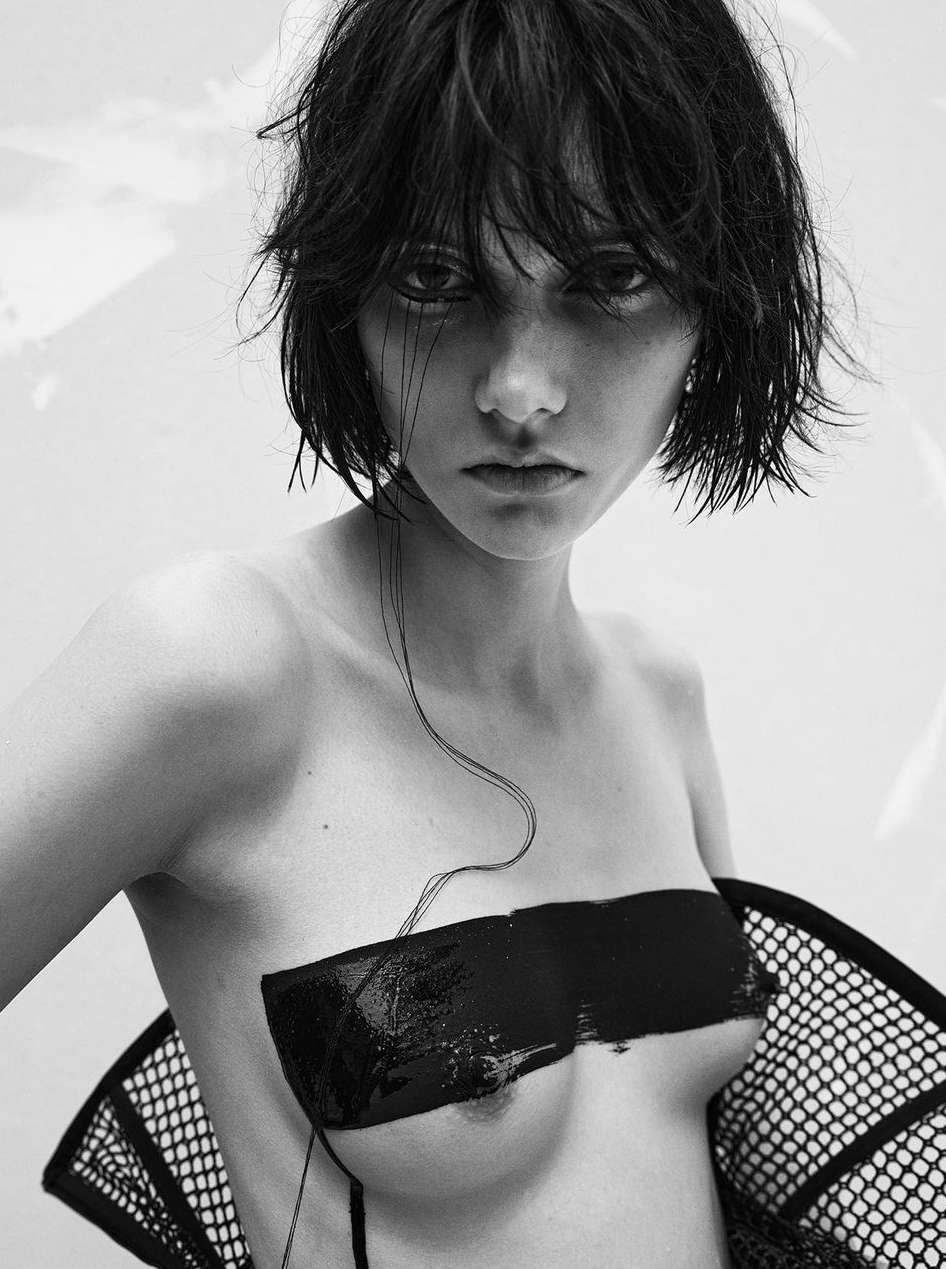 Striking black and white portrait of a young woman with a dark bob hairstyle, a blank black painted bar across her chest and fishnet details.