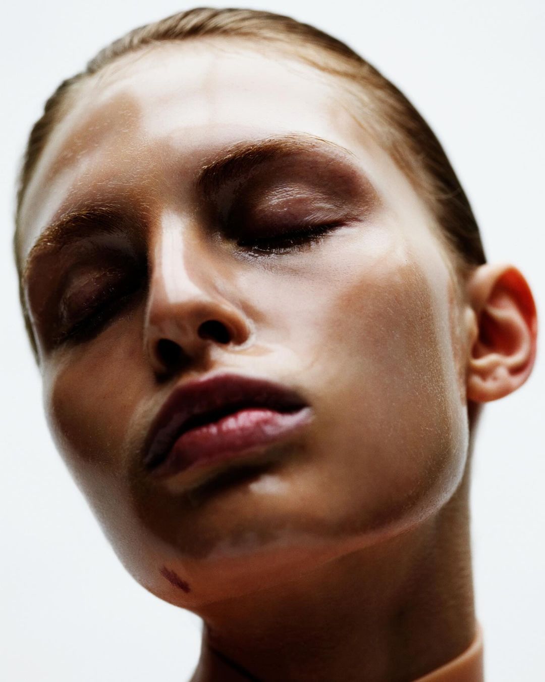 Close-up beauty shot of a young adult woman with her eyes closed, wet skin, and glossy red lips against a clean, bright, white background.