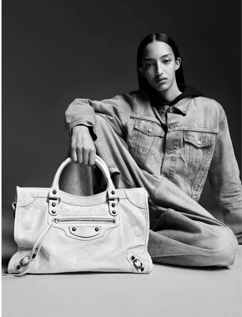 Monochrome studio shot of a woman in a denim jacket and jeans sitting with a light-colored leather handbag in a minimalist setting.