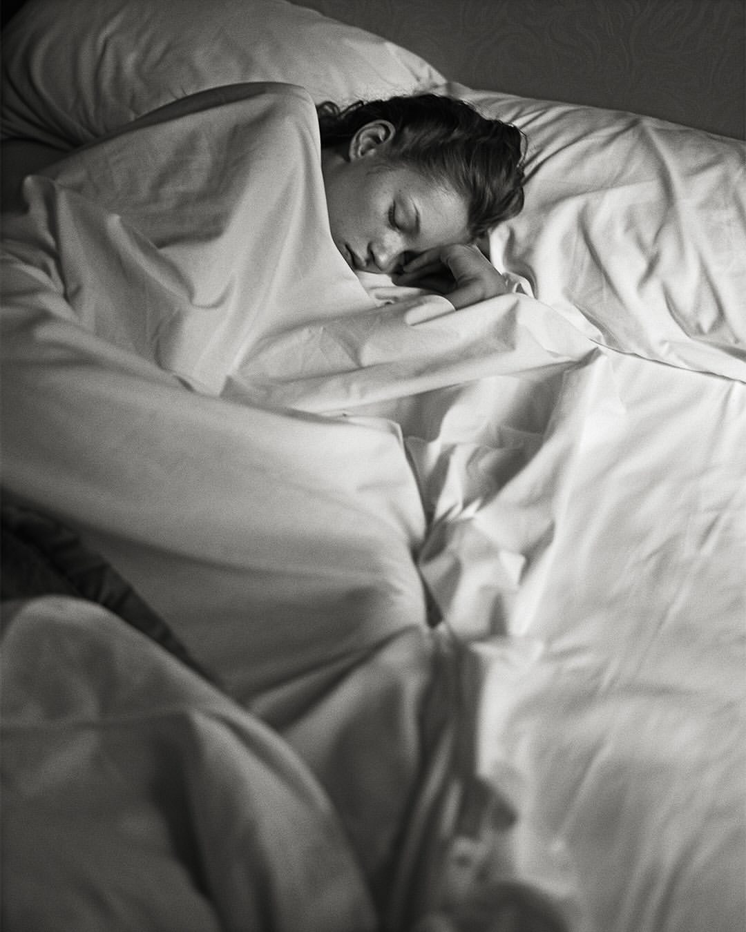 Woman sleeping peacefully in bed covered with a white sheet, resting her head on a pillow in a dimly lit room.