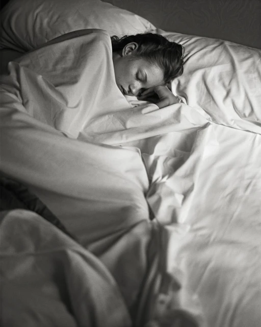 Woman sleeping peacefully in bed covered with a white sheet, resting her head on a pillow in a dimly lit room.