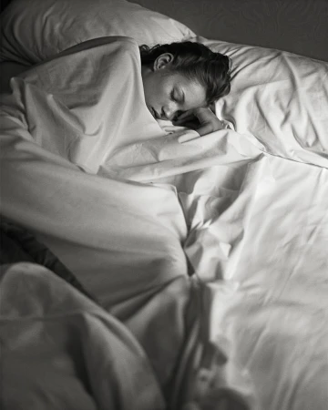 Woman sleeping peacefully in bed covered with a white sheet, resting her head on a pillow in a dimly lit room.