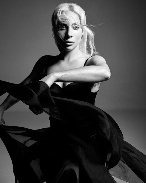 Black and white portrait of a woman with light hair wearing a black dress, striking a dynamic pose against a neutral background.