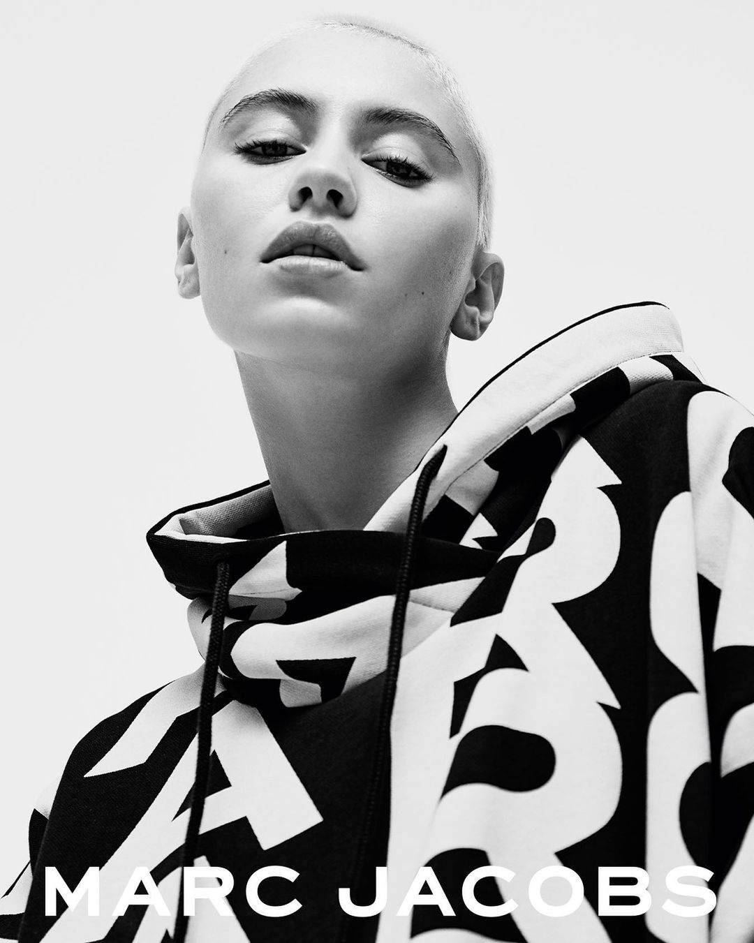 Close-up shot of a young adult female with a shaved head, wearing a black and white patterned hoodie with the Marc Jacobs logo.