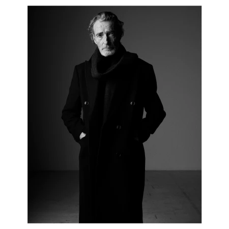 Portrait of an older man with grey hair wearing a black coat and scarf against a plain studio background.